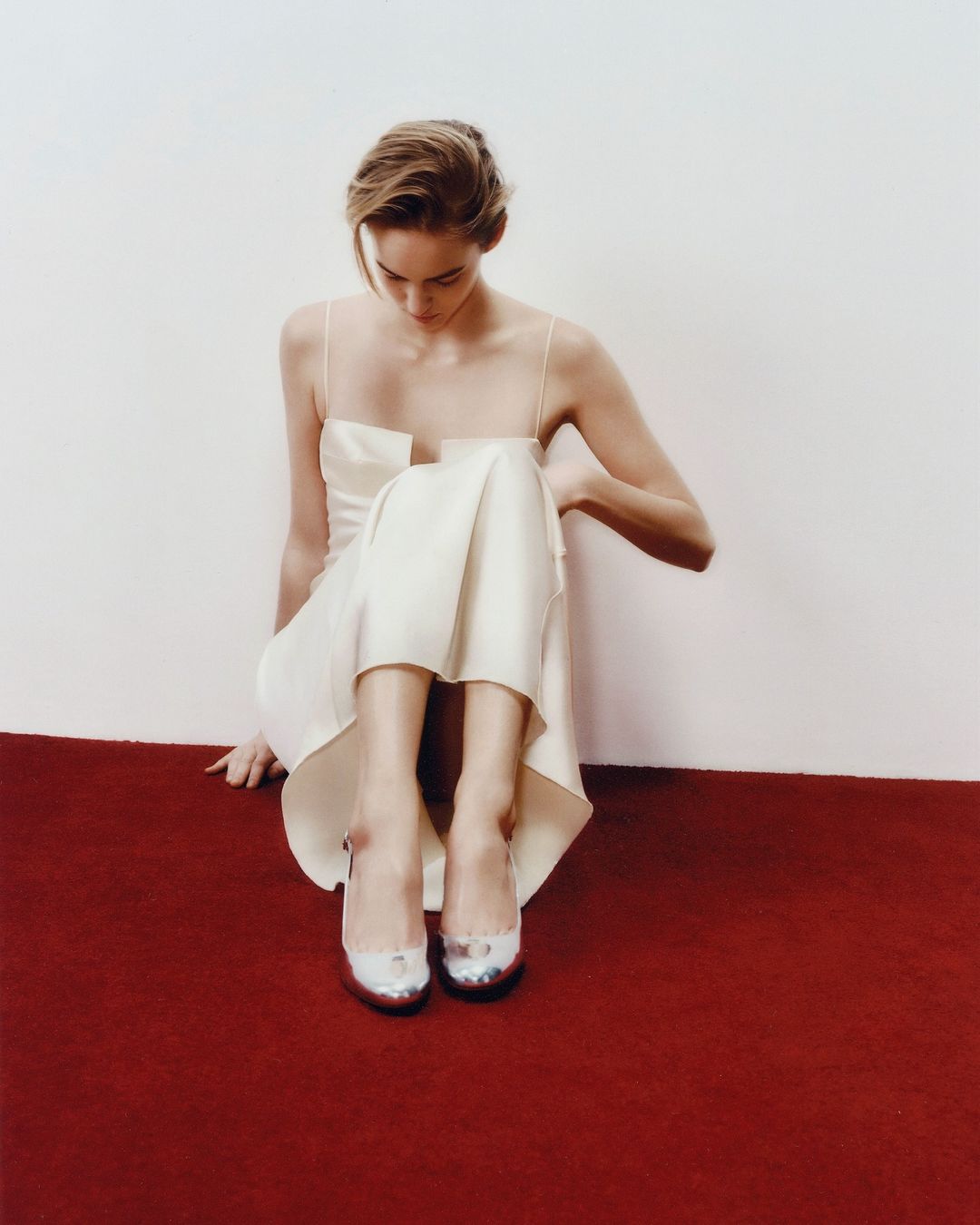 A blonde woman in a cream dress sits on a red rug with silver heeled shoes in a minimalist fashion studio shot with a white background.