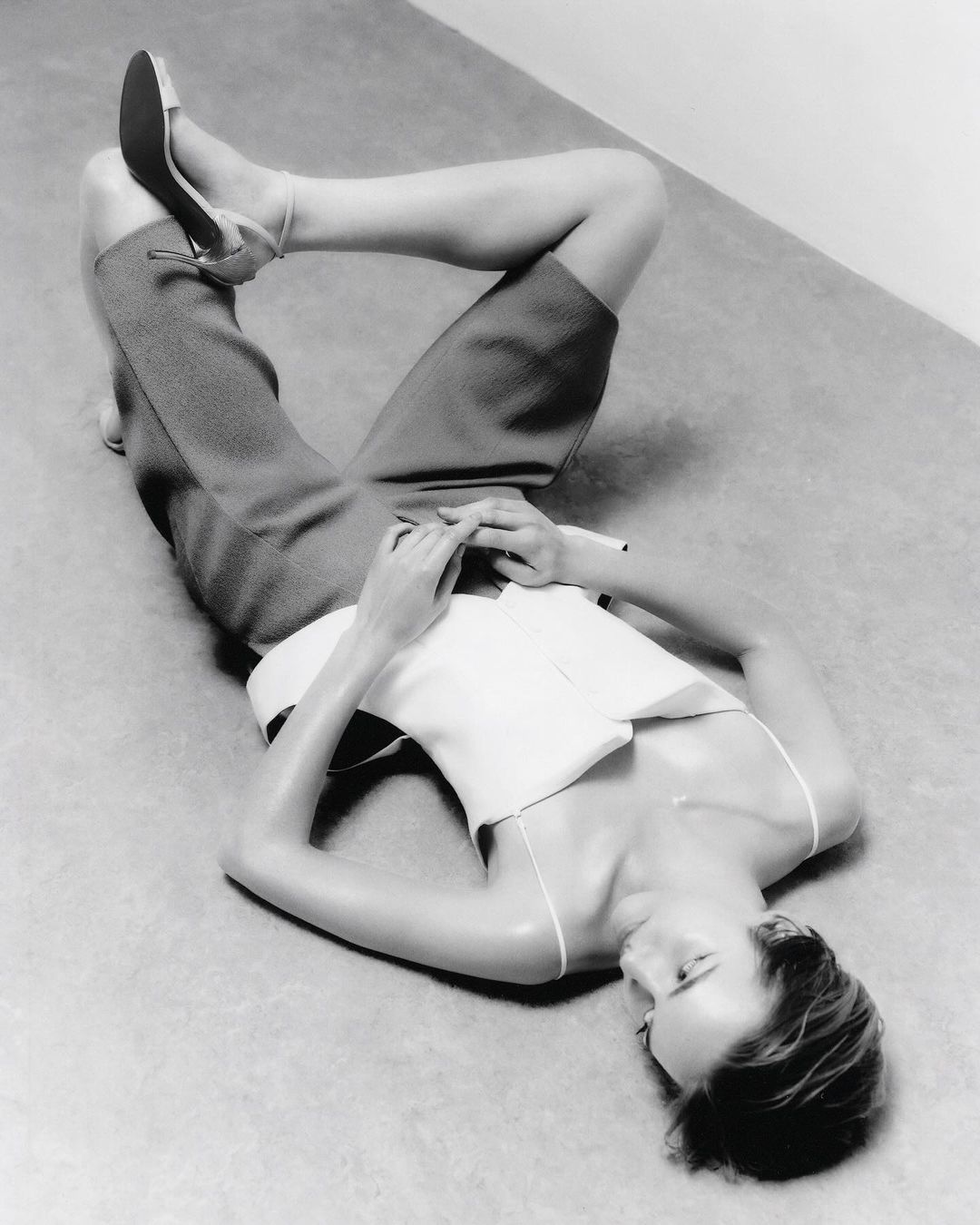 Monochrome portrait of a stylish woman lying on her back, wearing a white top, shorts, and heeled shoes in a minimalist studio setting.