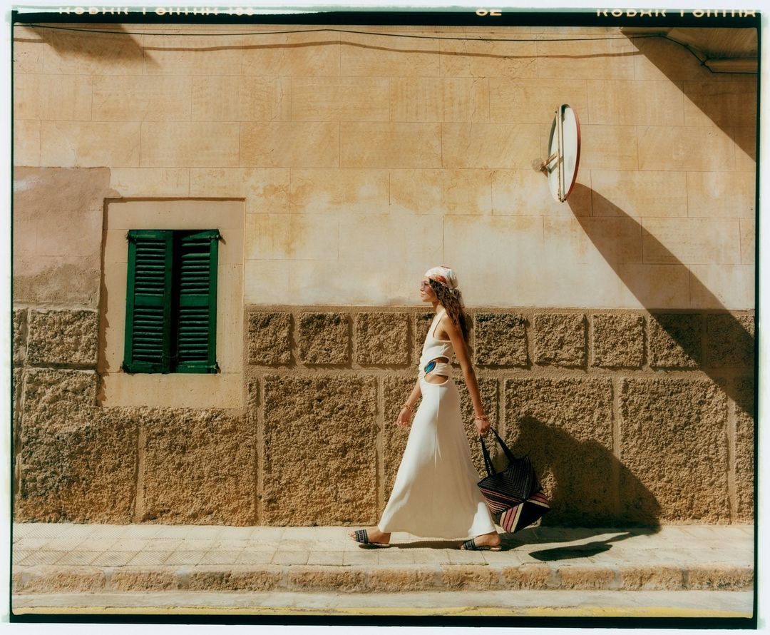 A woman in a white dress and headscarf walks along a sidewalk beside a textured tan building with a green-shuttered window and round mirror.