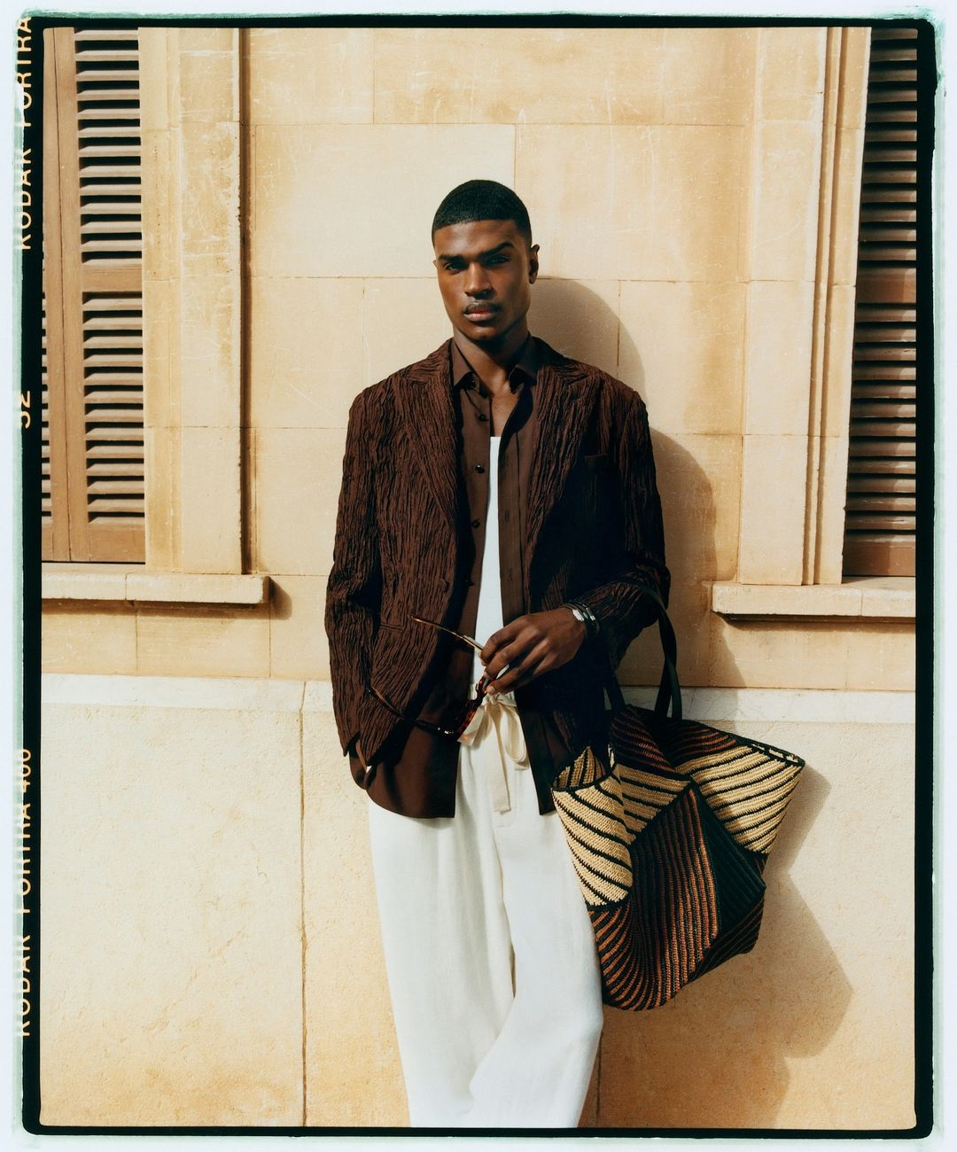 A dark-skinned male model poses against a cream wall near a window, wearing a brown jacket, shirt, and white pants, and holding sunglasses.