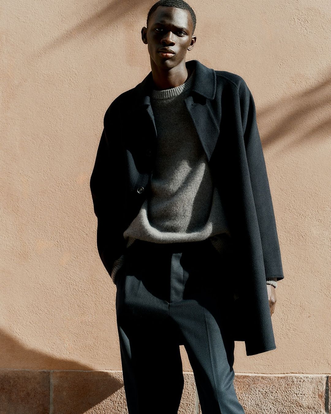 Portrait of a black male model wearing a grey sweater, dark trousers, and a black coat, posing against a neutral beige background.