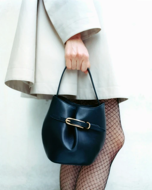 A woman in a cream coat holds a structured, dark teal bucket bag with a gold buckle, complemented by black fishnet stockings.