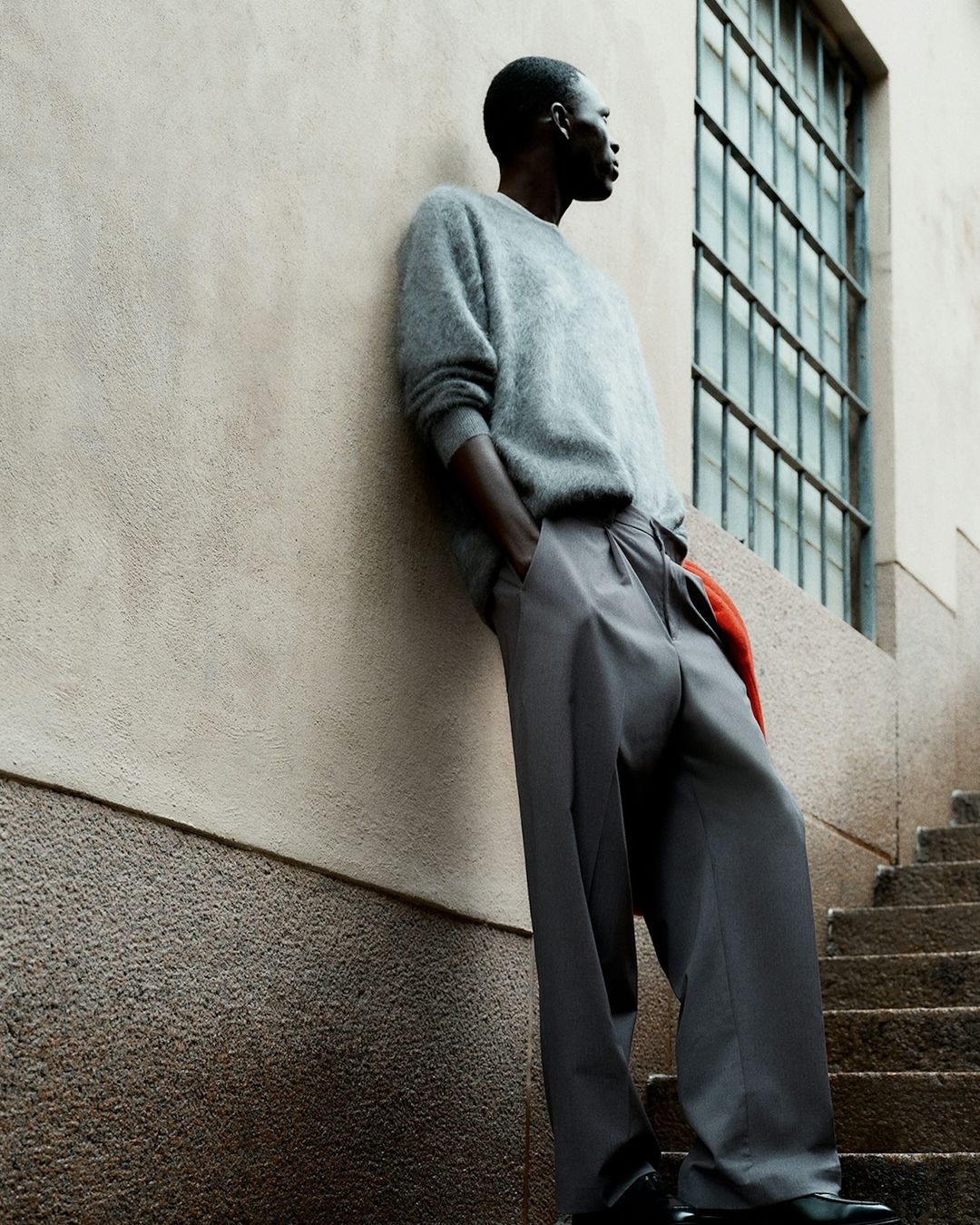 Side view of a Black male model in gray sweater and wide-leg trousers, leaning against a wall next to a window, with stairs at the base.