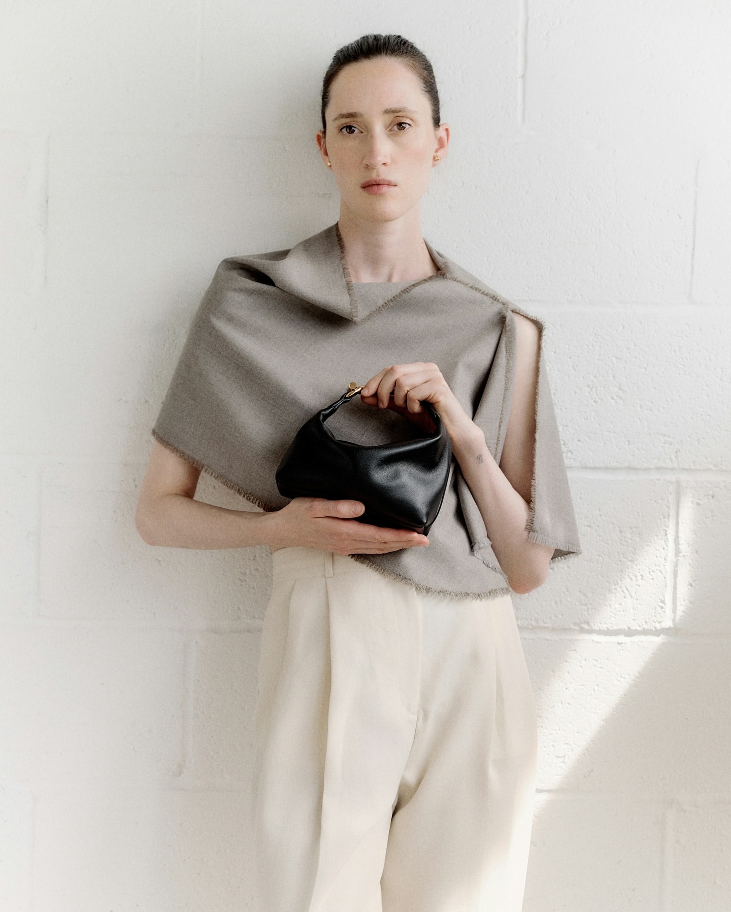 Fashion shot of an adult female model holding a black leather handbag in front of a textured white wall, wearing beige slacks.