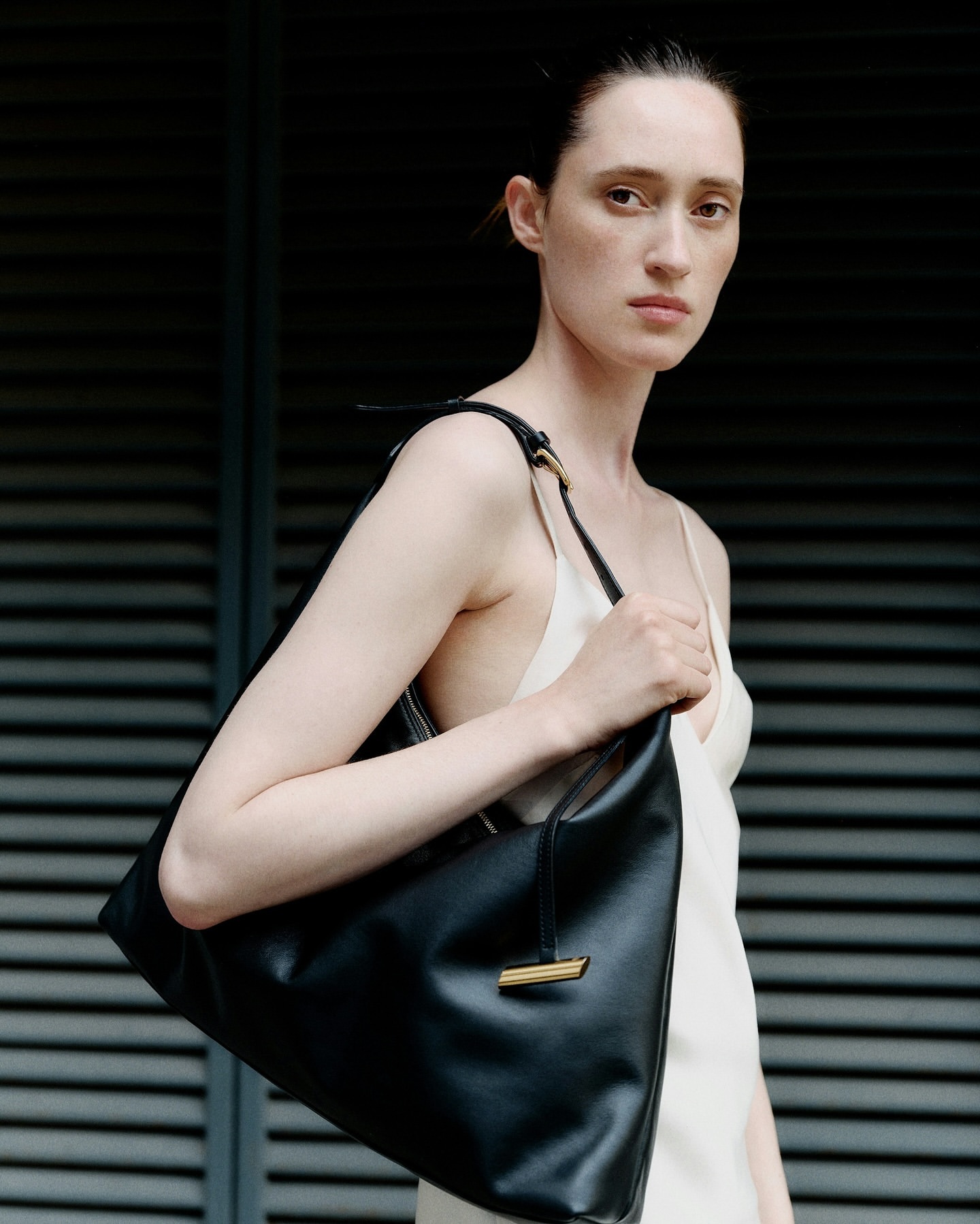 Fashion portrait of a slender woman with a black leather handbag, standing in front of a gray louvered wall, minimalist composition.