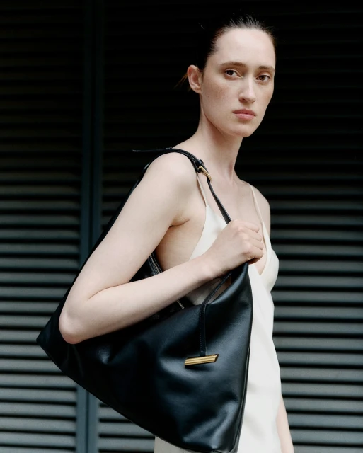 Fashion portrait of a slender woman with a black leather handbag, standing in front of a gray louvered wall, minimalist composition.