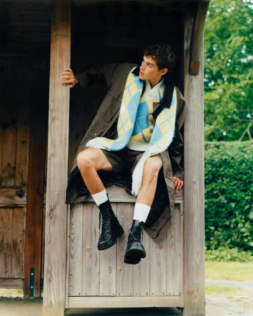 Young man in shorts, sweater, trench coat, socks and boots, sits in a wooden bus shelter, green trees visible in background, medium shot.