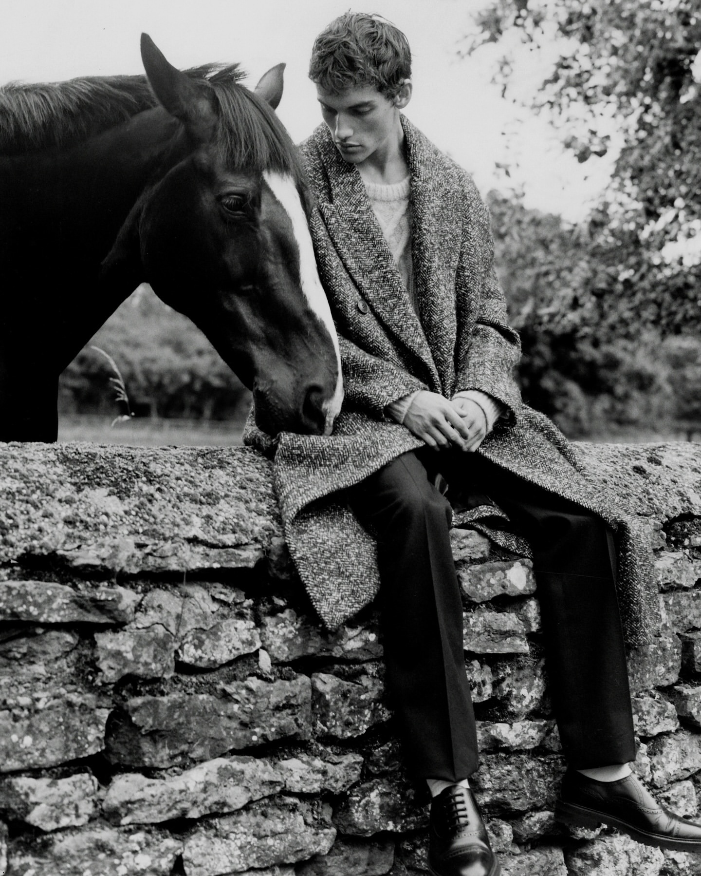 Black and white shot of a young man in a coat sitting on a stone wall next to a horse in a rural setting with trees in the background.