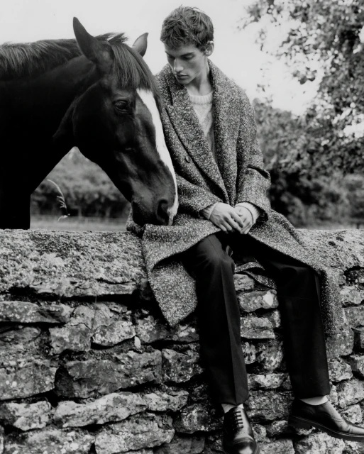 Black and white shot of a young man in a coat sitting on a stone wall next to a horse in a rural setting with trees in the background.