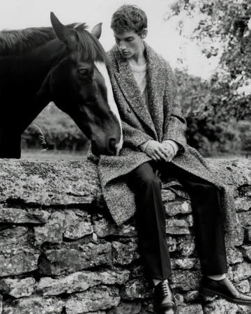 Black and white shot of a young man in a coat sitting on a stone wall next to a horse in a rural setting with trees in the background.