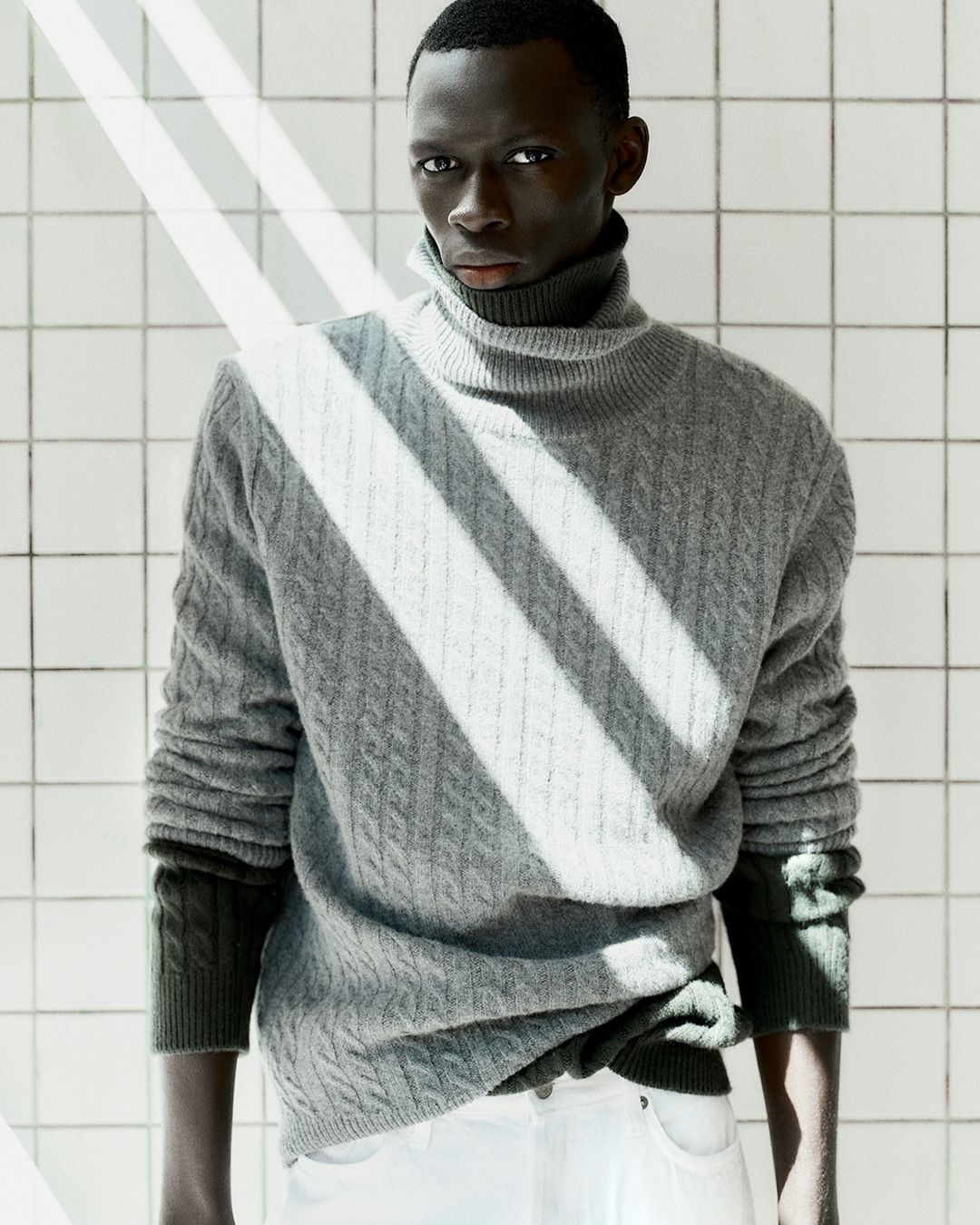 Black man wearing a grey cable-knit turtleneck sweater against a white tiled wall background