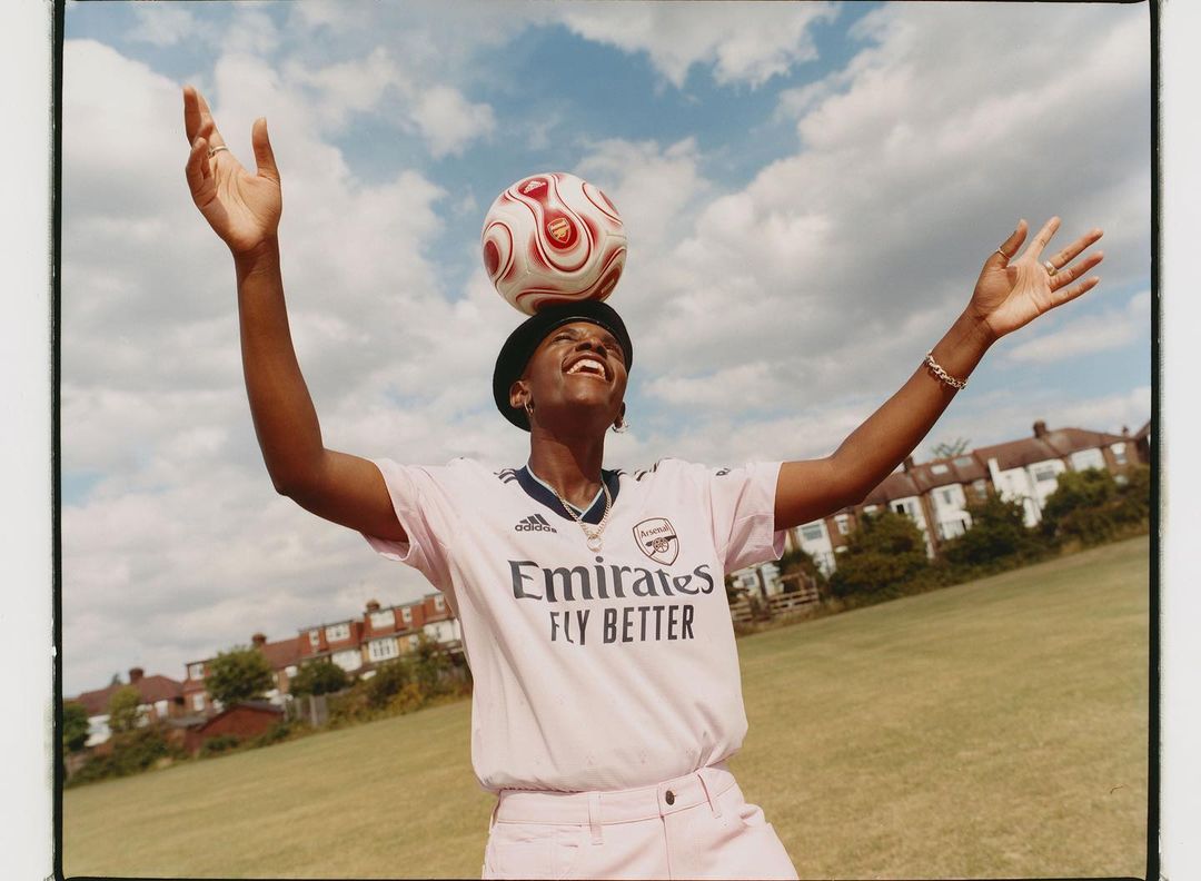 Female soccer player balances a soccer ball on her head in an outdoor setting with residential buildings and a cloudy sky in the background.