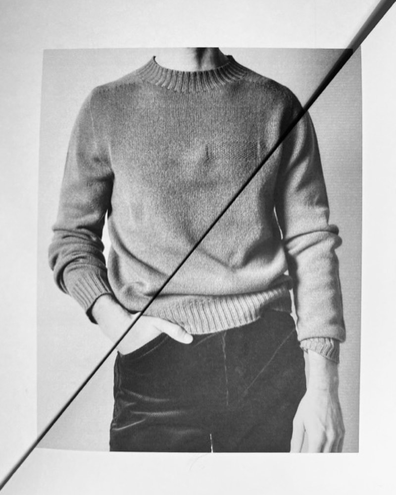 Monochrome photograph of a torso in a textured sweater with hand in pocket, bisected by a strong diagonal line against a neutral backdrop.