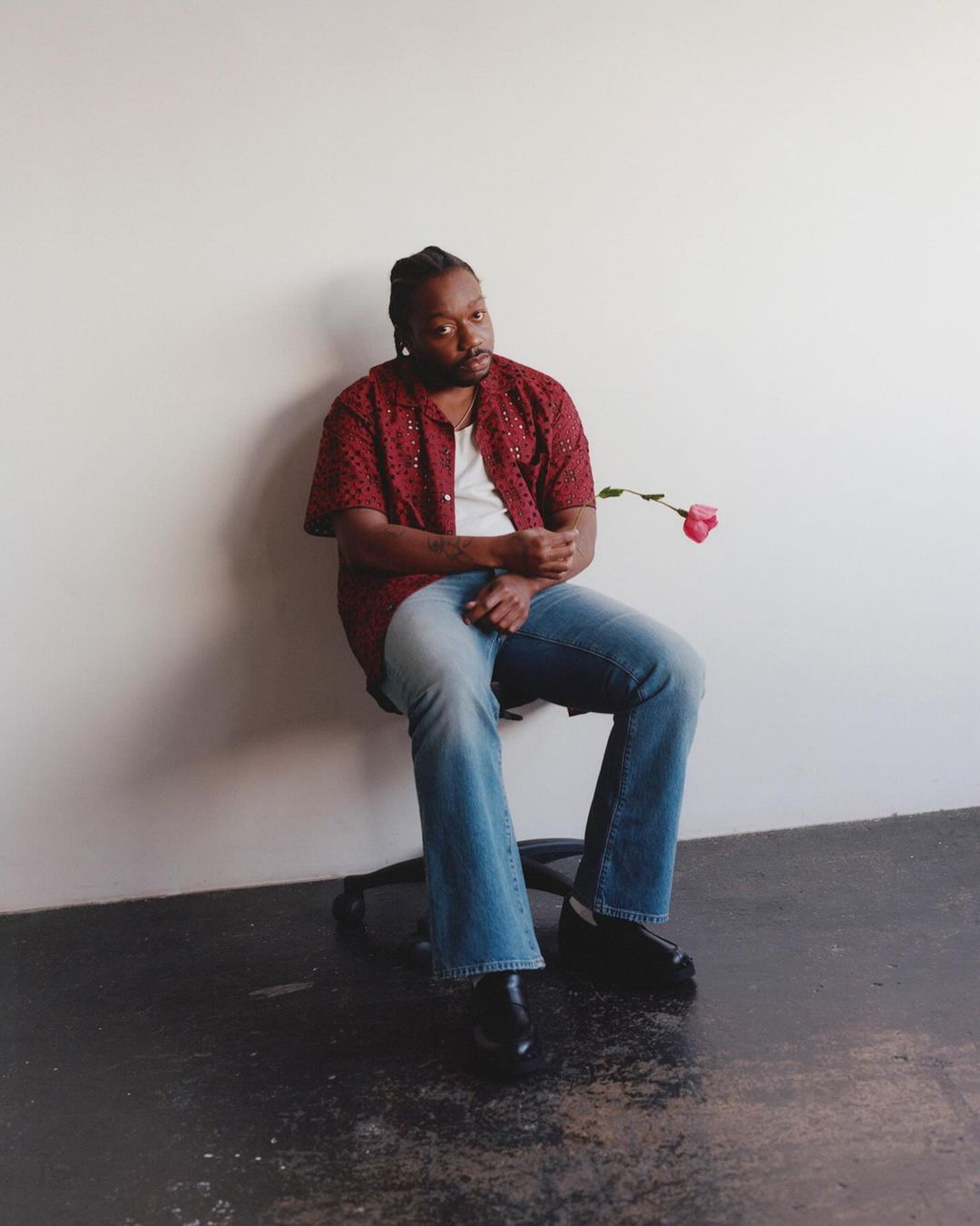 Adult male seated with rose in photo studio: man in printed shirt and jeans sits on stool against wall, holding a single rose.