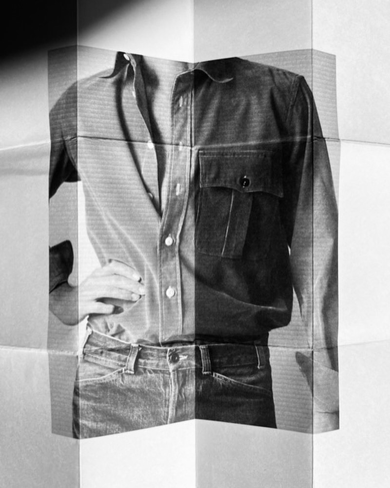 Monochromatic image of model wearing a collared shirt, jeans, hand on hip, presented on folded paper; evokes a vintage editorial style.