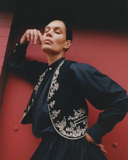 Woman posing in dark blue blouse and embroidered vest against a red brick wall with head tilted and hand near face.