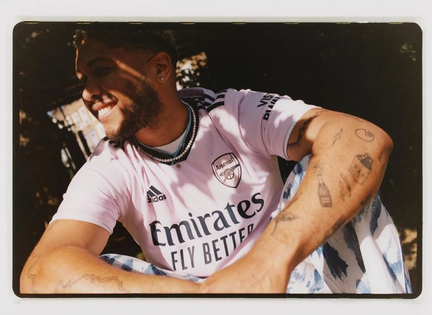 Smiling man in pink soccer jersey with Arsenal logo, patterned pants, wearing pearl necklace, arm tattoos, in natural light.