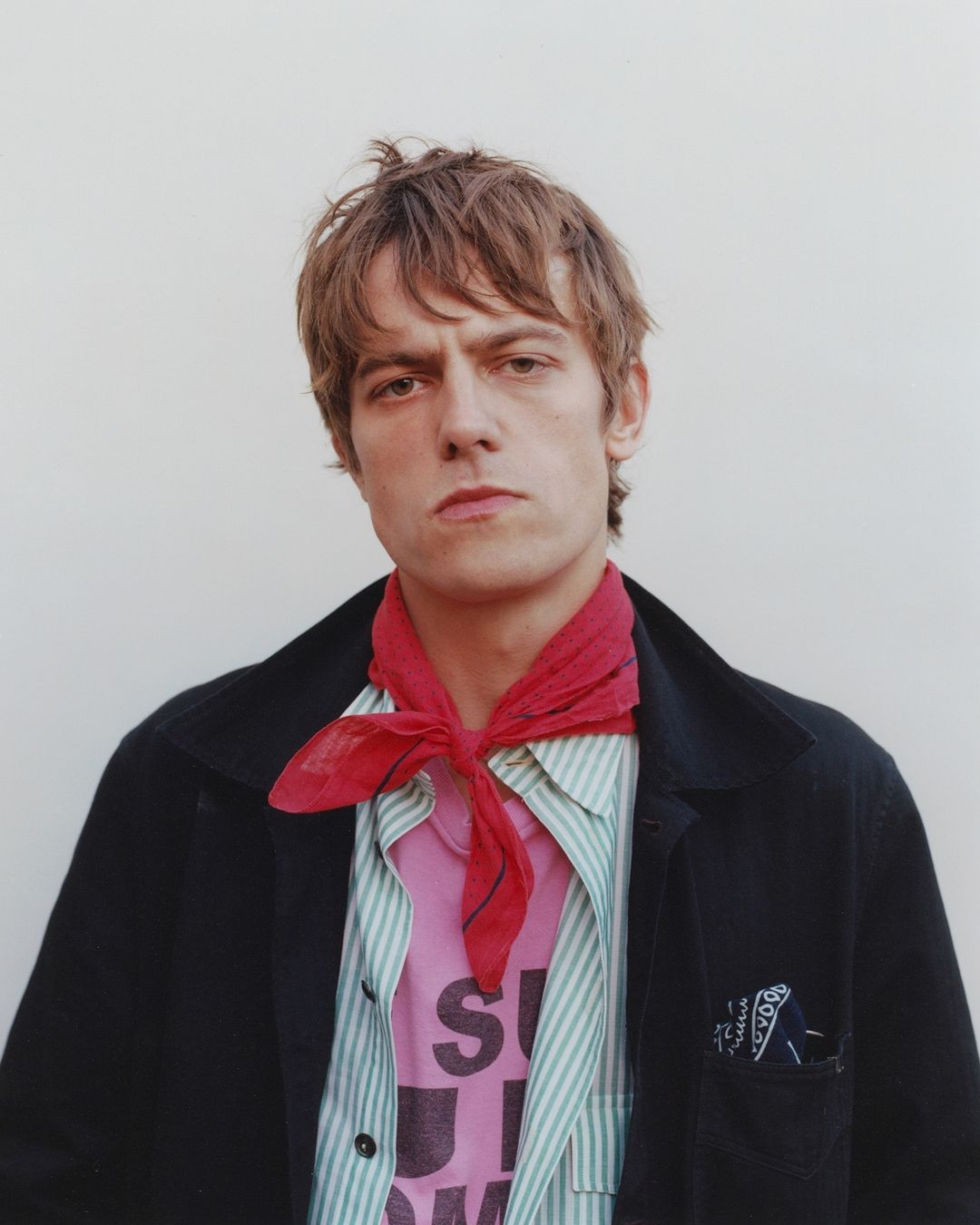 Portrait of a young adult man with brown hair wearing a red scarf, pink T-shirt, and black jacket against a neutral background.