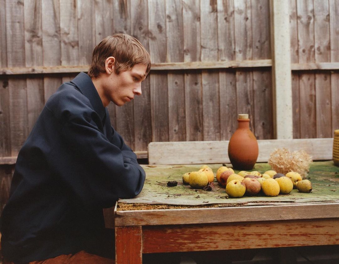 Adult man sits at a wooden table with fresh fruit in an outdoor setting near a weathered wood fence, wearing a dark blue jacket.