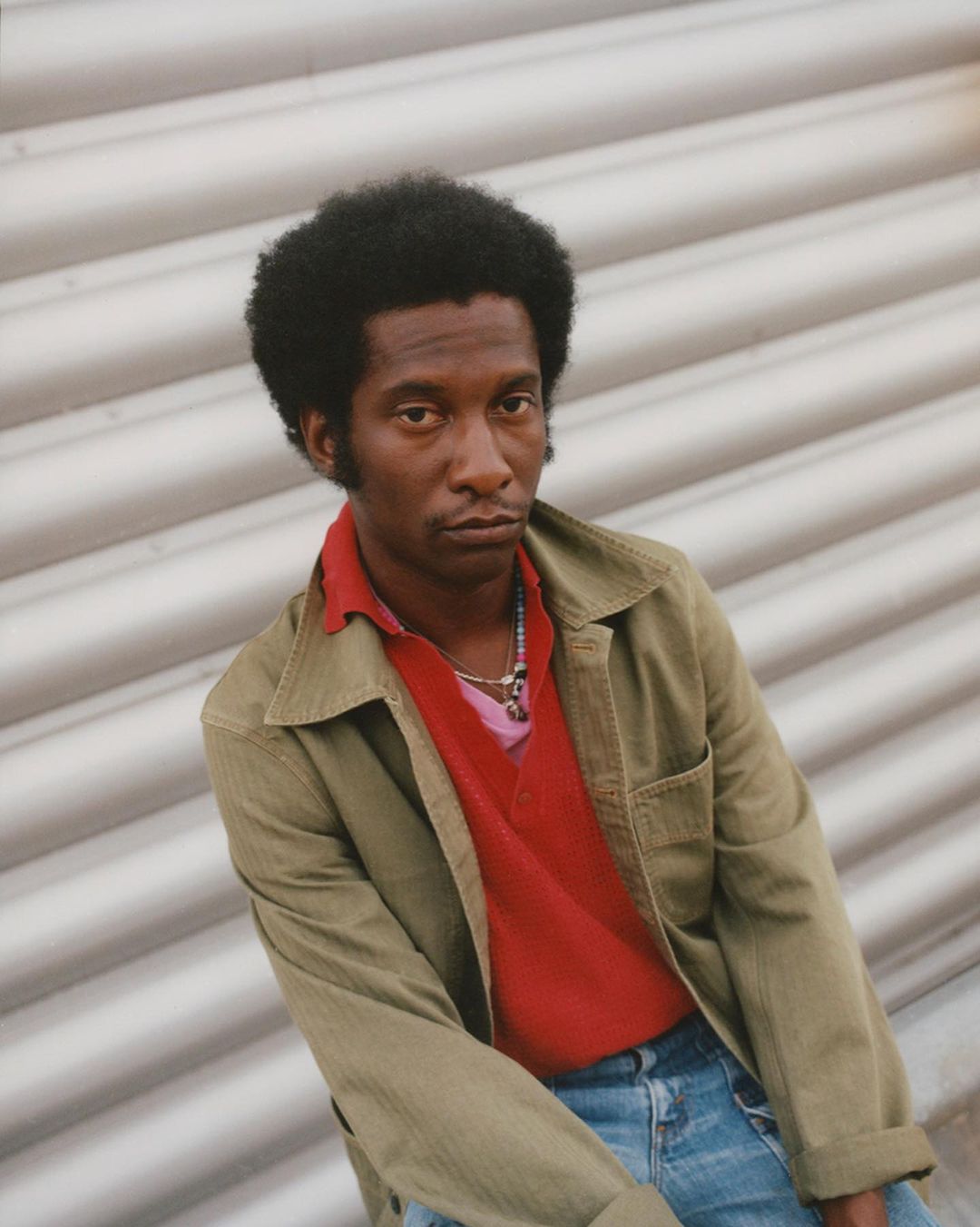 Portrait of a dark-skinned adult male with an afro, wearing a tan jacket over a red polo shirt, in front of a textured gray wall.