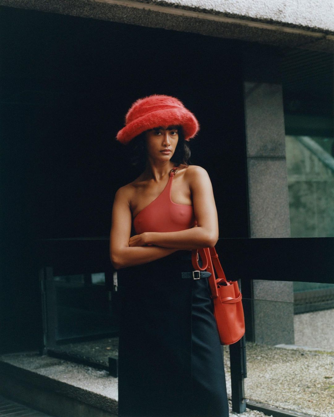 A woman poses in a salmon-colored one-shoulder top and dark skirt with a matching fuzzy hat and shoulder bag in an outdoor setting.
