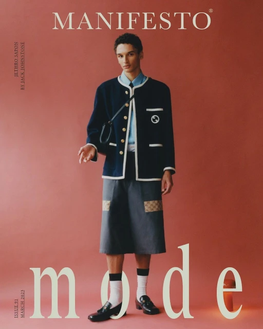 Fashion magazine cover featuring a slender young man in a blue jacket, skirt, and black loafers on a pink backdrop with large text.