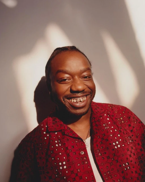 Close-up studio shot of a smiling adult male with a gold grill, wearing a patterned red collared shirt against a light wall.