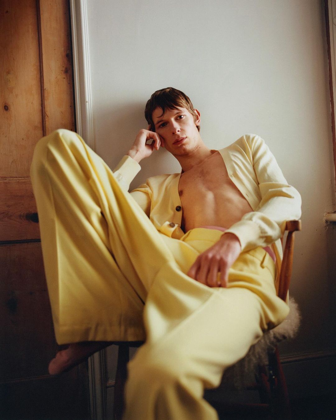 Young adult man in yellow cardigan and pants sits in chair. Indoor portrait with a relaxed pose and neutral facial expression.