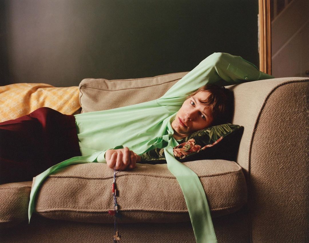Young man rests on a beige couch wearing a light green shirt and red pants. The setting is indoors with a muted green wall.