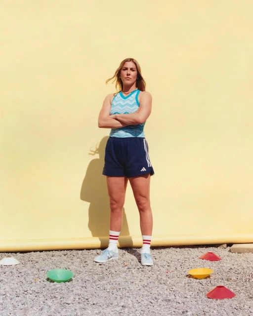 A female athlete stands with arms crossed in blue athletic shorts and patterned tank top against a yellow wall, practicing on a gravel surface.