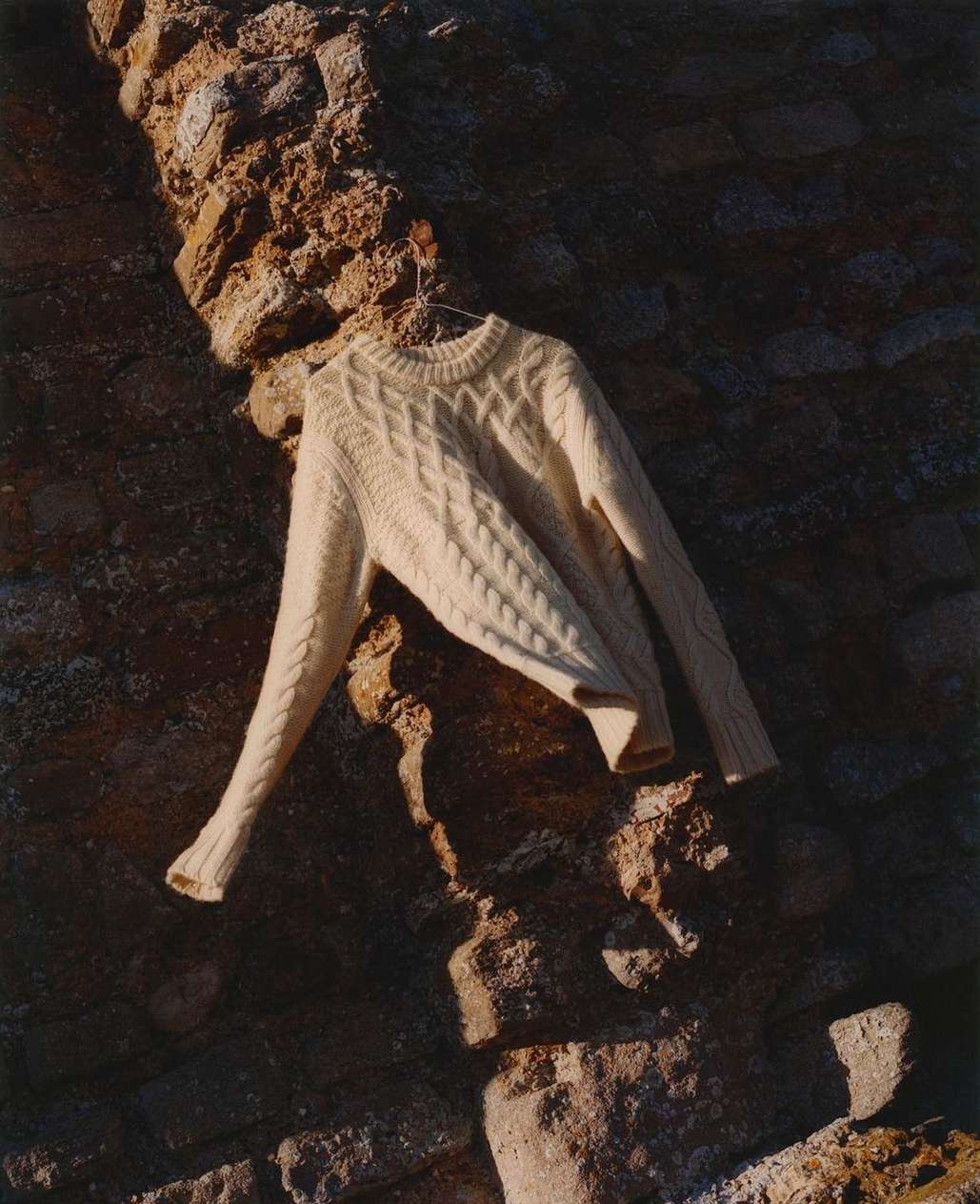 Cream-colored cable knit sweater hanging on a metal hanger against a textured stone wall, casting shadows in a minimalist fashion shot.