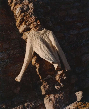 Cream-colored cable knit sweater hanging on a metal hanger against a textured stone wall, casting shadows in a minimalist fashion shot.