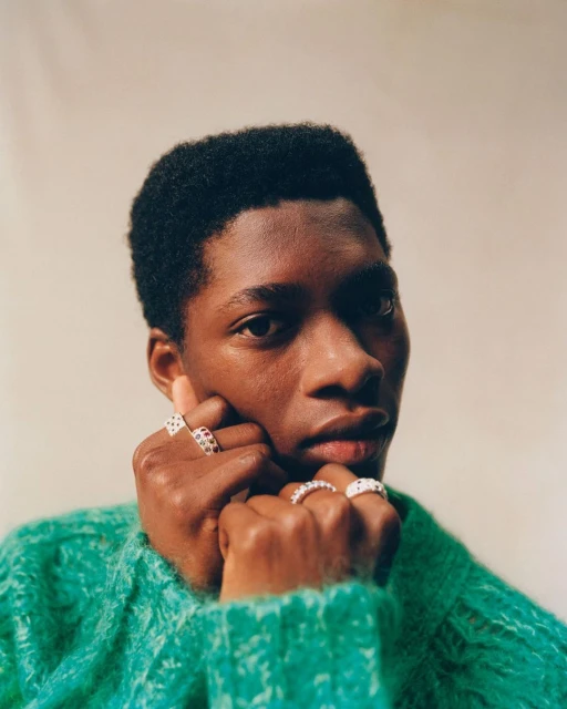 Portrait of a Black man wearing a green sweater and multiple rings on his fingers, close-up shot against a neutral background.