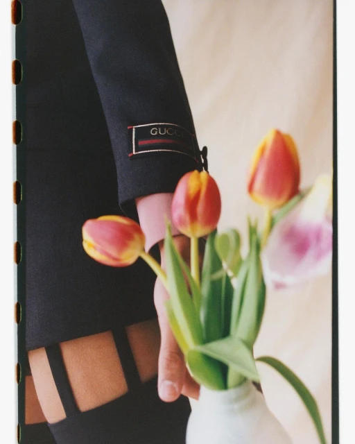 Close-up of a black jacket sleeve with Gucci logo, a hand, and a vase of red, yellow, and pink tulips, indoors, soft lighting.