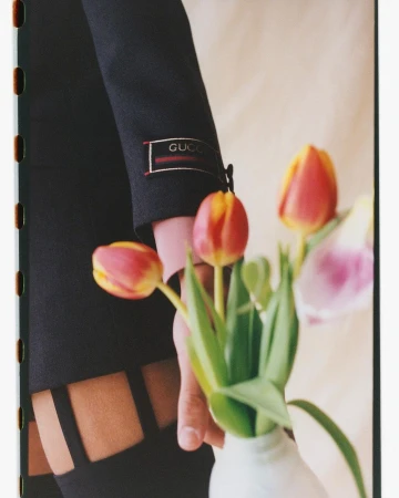 Close-up of a black jacket sleeve with Gucci logo, a hand, and a vase of red, yellow, and pink tulips, indoors, soft lighting.