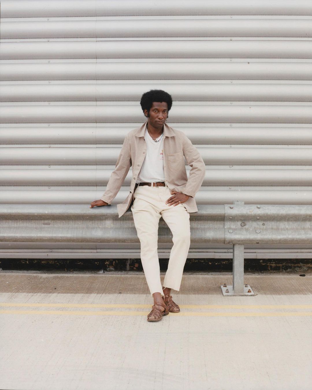 Stylish adult male poses against a textured silver wall wearing beige jacket, white shirt, and brown sandals, exuding confidence.