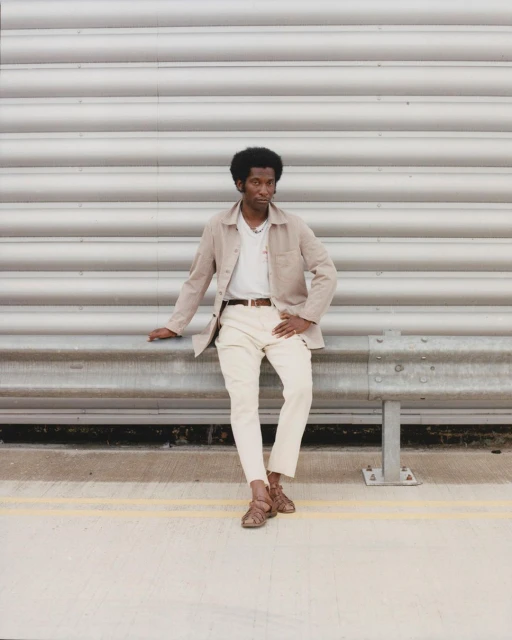 Stylish adult male poses against a textured silver wall wearing beige jacket, white shirt, and brown sandals, exuding confidence.