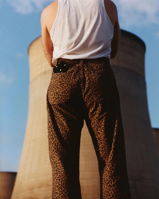A person wearing leopard print pants and a white tank top stands in front of cooling towers against a blue sky on a sunny day.