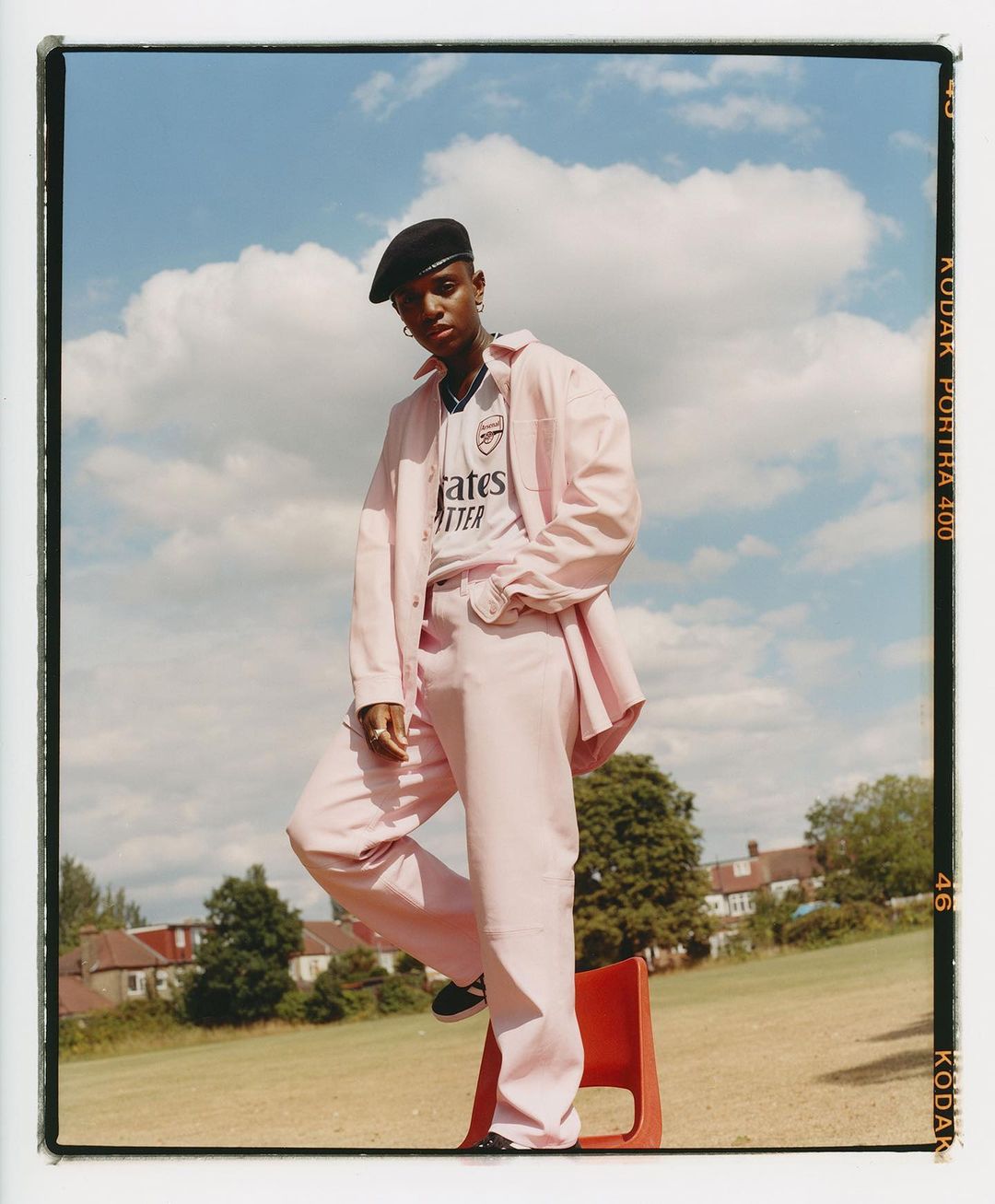 Young adult female in a pink suit and beret poses outdoors with a football jersey, red chair, houses, and blue sky with clouds.