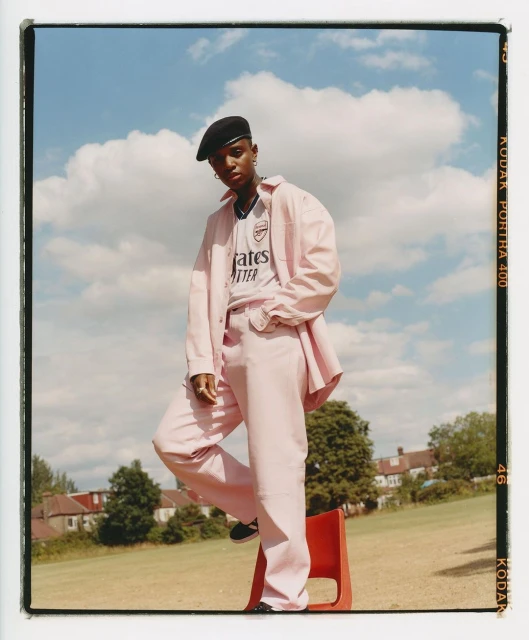 Young adult female in a pink suit and beret poses outdoors with a football jersey, red chair, houses, and blue sky with clouds.