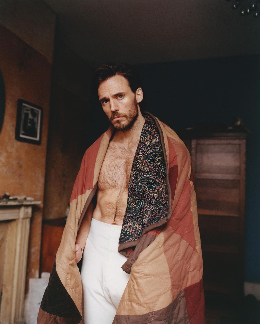 Indoor portrait of adult male with beard, wrapped in colorful quilt, wearing white underwear. Relaxed pose, natural light, interior background.