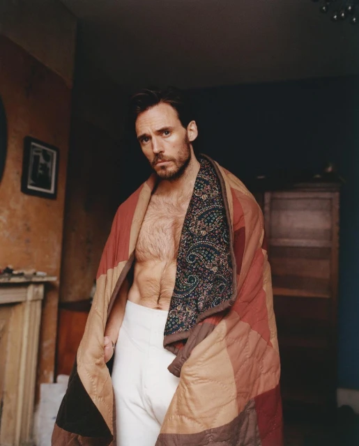 Indoor portrait of adult male with beard, wrapped in colorful quilt, wearing white underwear. Relaxed pose, natural light, interior background.
