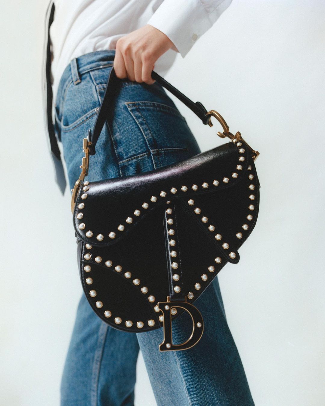 Close-up of a black leather saddle bag with pearl embellishments, held by an adult female wearing a white shirt and jeans.