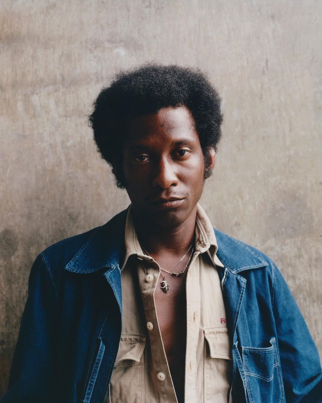 Portrait of an African-American male in a denim jacket and tan shirt, posing in front of a textured grey wall. The shot is medium-range.