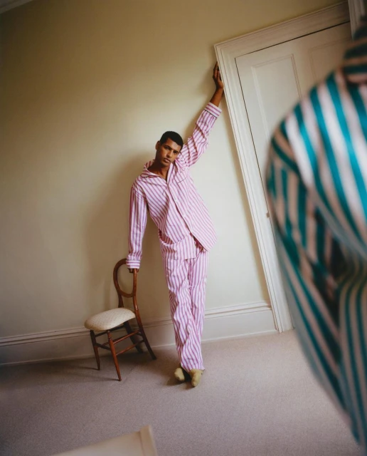 A person wearing pink and white striped pajamas leaning against a white door with a chair in an indoor setting, captured in a medium shot.