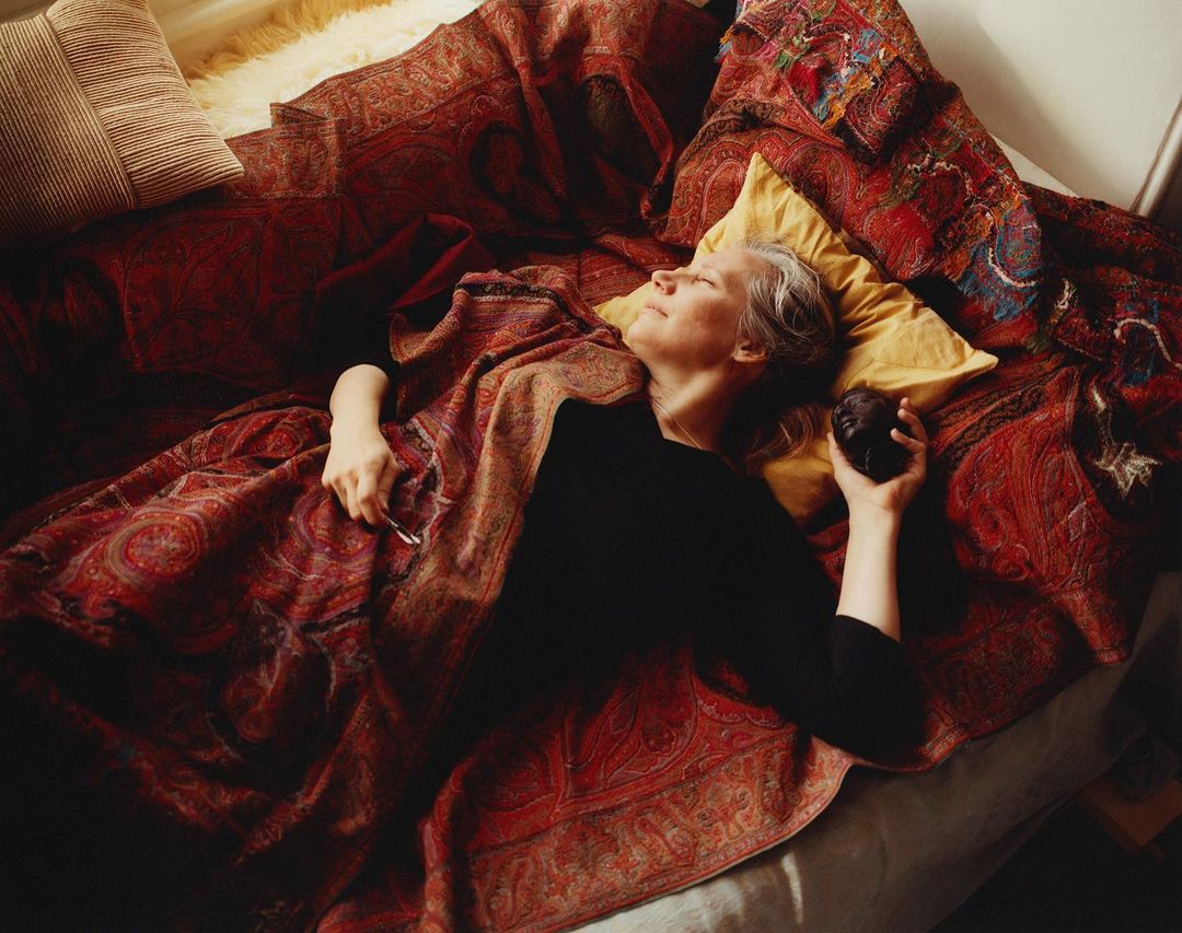 Adult woman with fair skin and gray hair is reclining on a paisley blanket on a bed, holding a dark object, with a neutral facial expression.