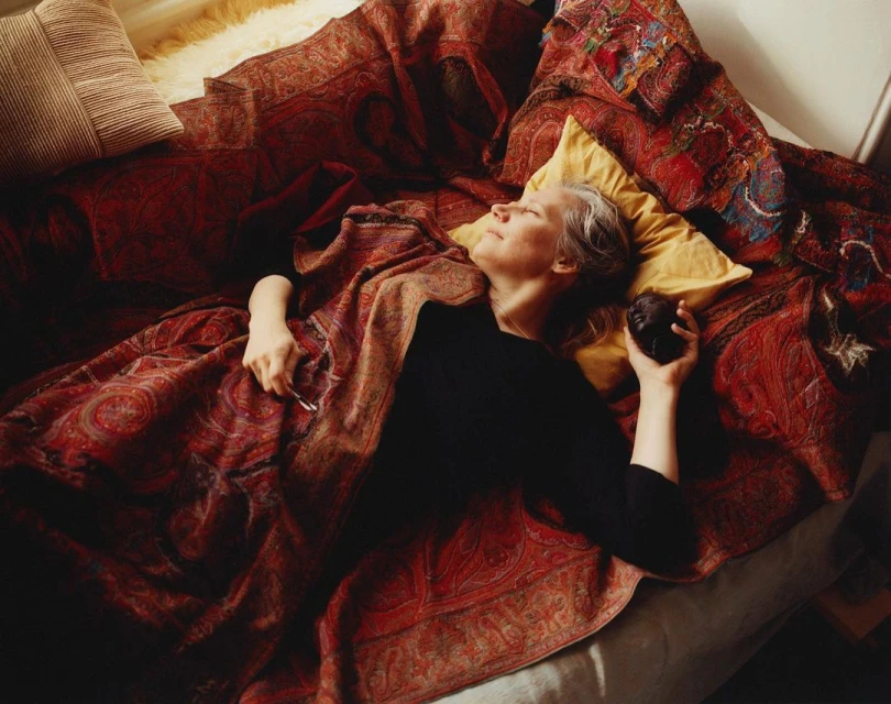 Adult woman with fair skin and gray hair is reclining on a paisley blanket on a bed, holding a dark object, with a neutral facial expression.