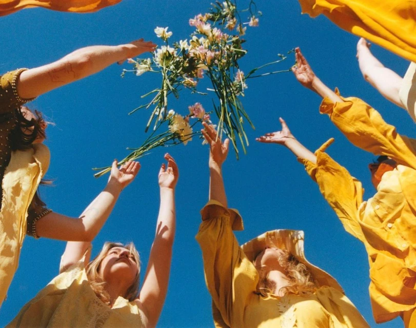 Low-angle shot of group reaching for bouquet of flowers against a clear blue sky, wearing yellow clothing, captured with natural lighting.