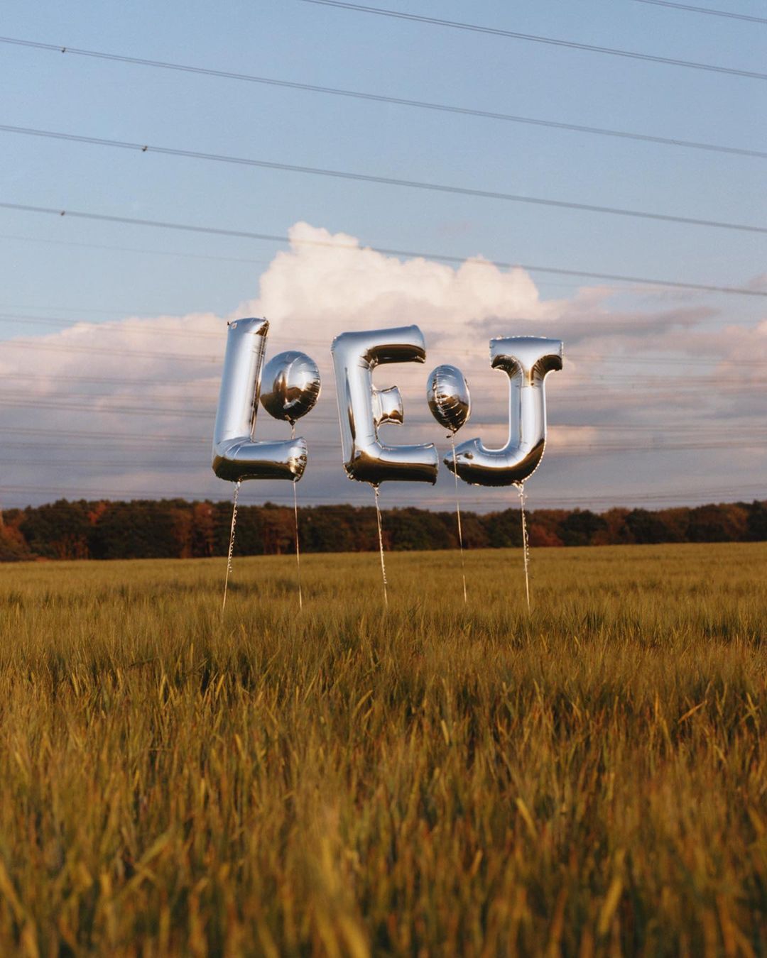 Shiny silver balloon letters spelling out 'LEJ' float above a golden wheat field under a cloudy blue sky, framed by distant trees.