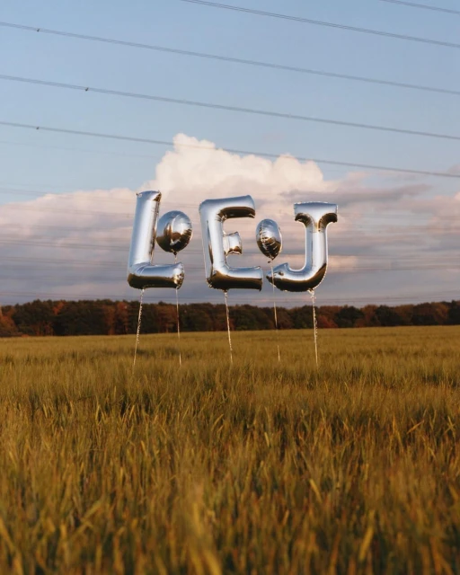 Shiny silver balloon letters spelling out 'LEJ' float above a golden wheat field under a cloudy blue sky, framed by distant trees.